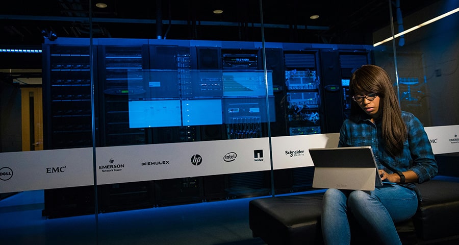 A young IT technician works on her laptop in front of a data center